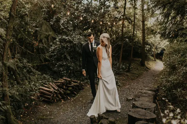 Bride and groom walking on tree-lined path with string lights in wooded Washington wedding setting. Lovely Events.
