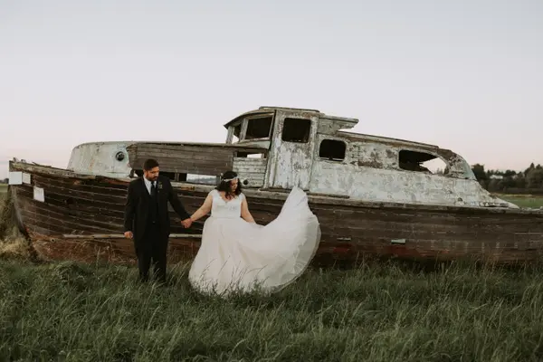 Bride and groom in grassy field beside weathered boat at outdoor Pacific Northwest wedding. Lovely Events.