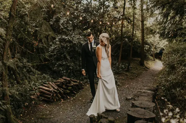 Bride and groom walking on tree-lined path with string lights in wooded Washington wedding setting. Lovely Events.
